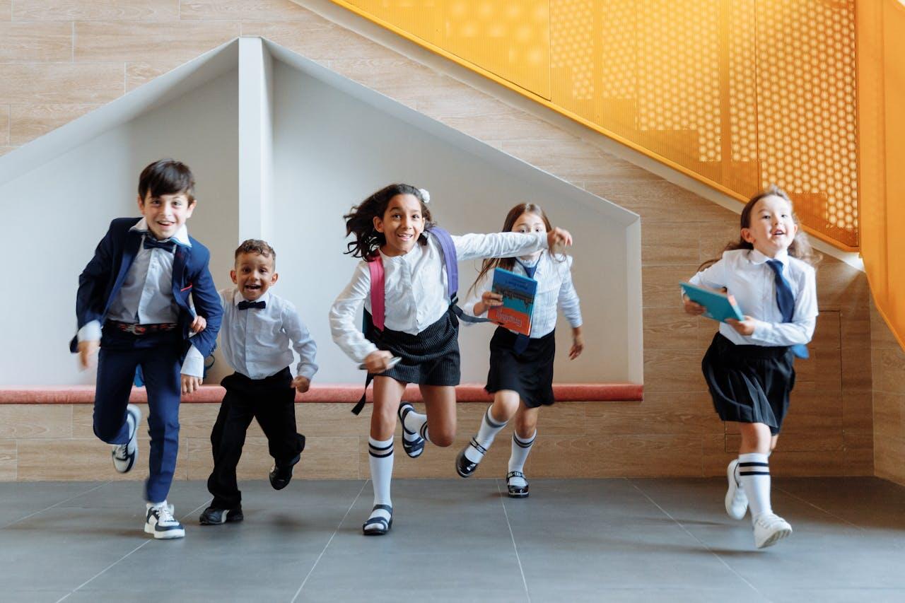 A group of children in formal attire joyfully runs down a corridor, holding books with bright orange accents on the staircase.