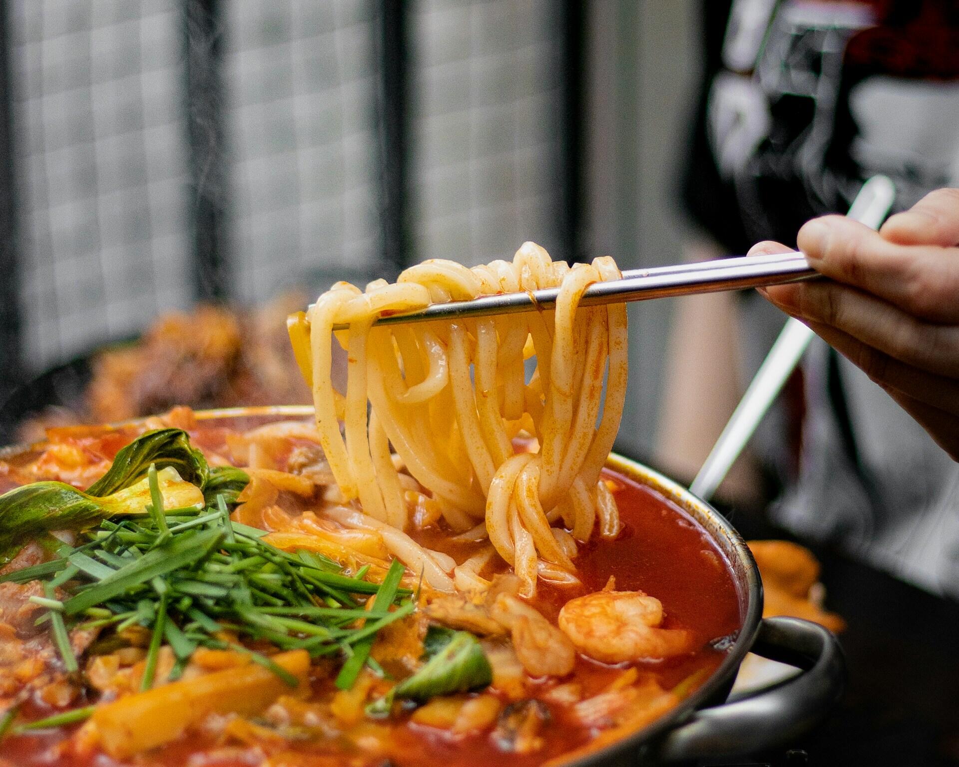 A person holding aloft noodles from a spicy soup pot.