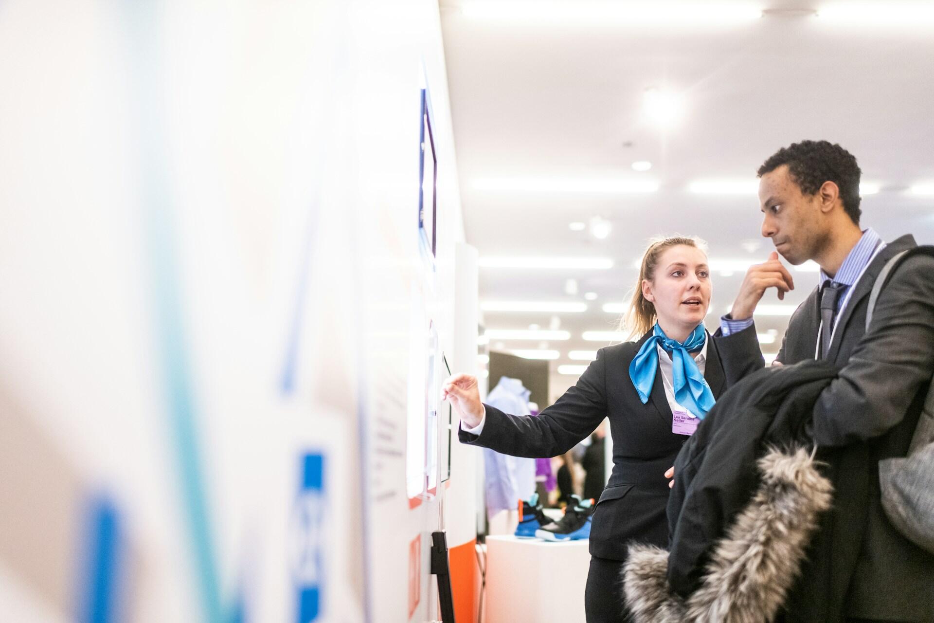 A professional woman gestures while explaining information to a man at a conference, surrounded by displays and attendees.