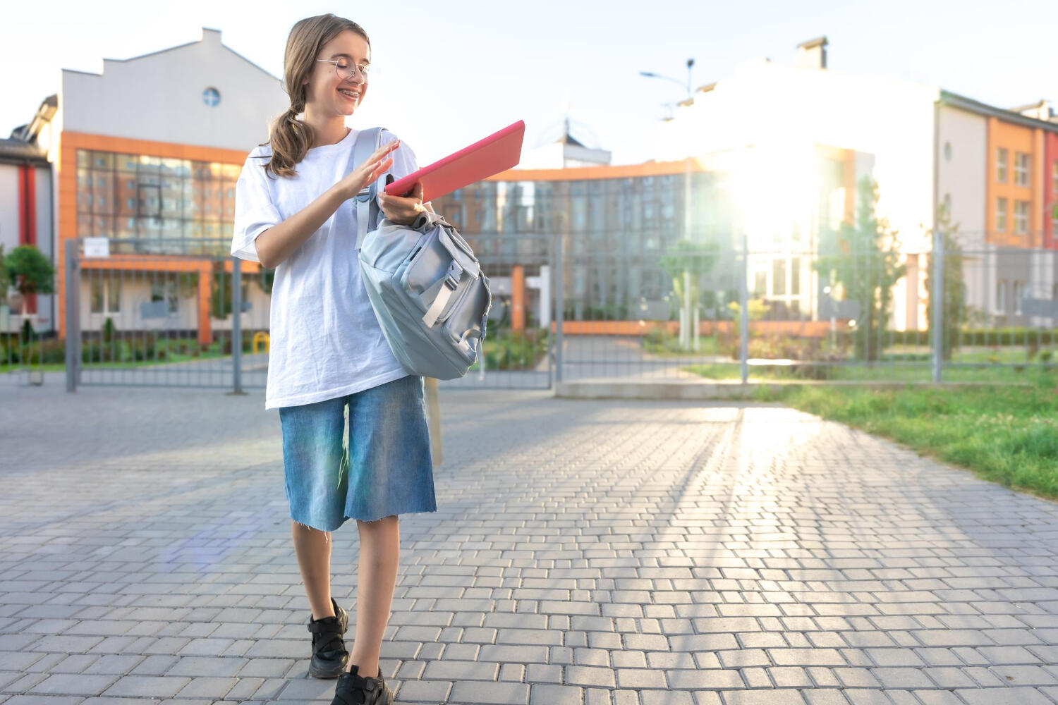 A student in a white t-shirt and denim shorts walks towards a school, holding a pink notebook and wearing a backpack.