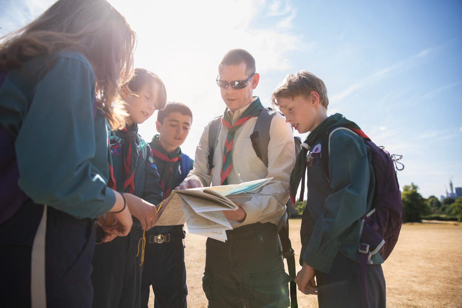 A group of scouts gathers outdoors, examining a map and discussing their next adventure under a sunny blue sky.