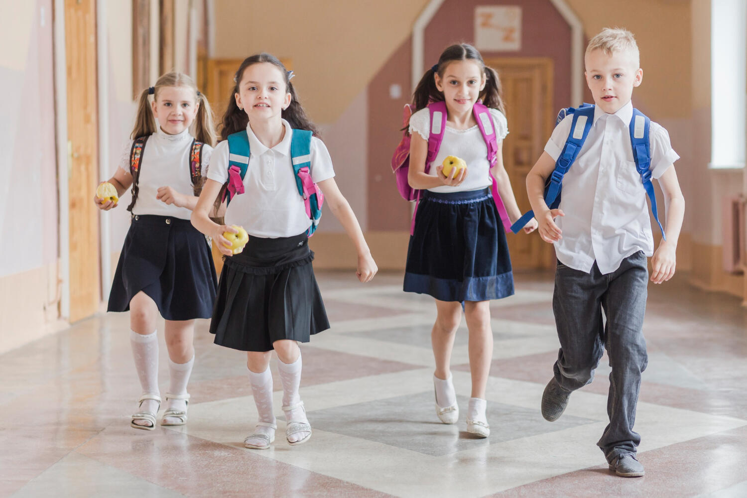 Four schoolchildren wearing backpacks and uniforms walk down a brightly lit hallway, each holding an apple.