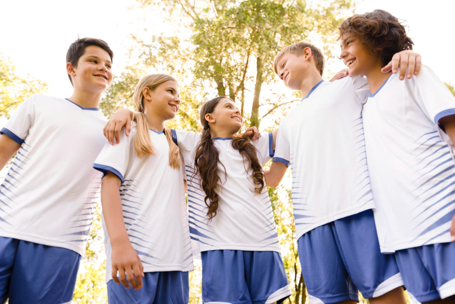 A group of five young athletes in sports uniforms stand closely together, smiling and enjoying a sunny day outdoors.