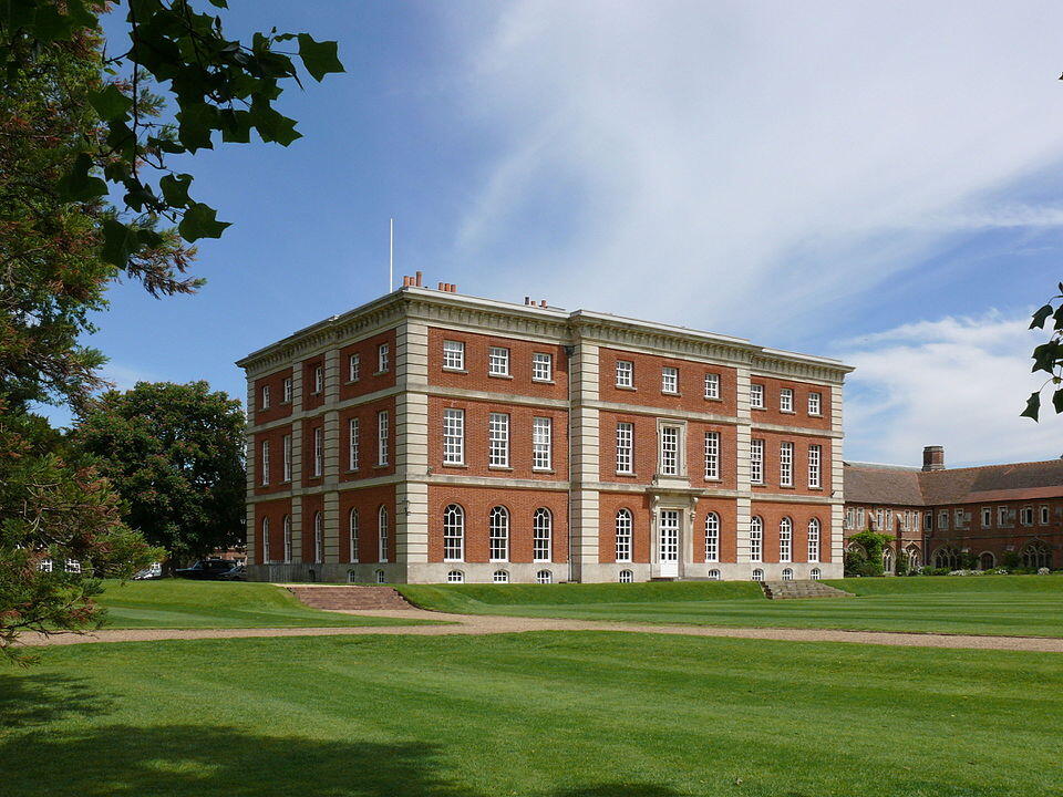 A large, two-story red brick building with white trim, surrounded by green grass and trees under a partly cloudy sky.