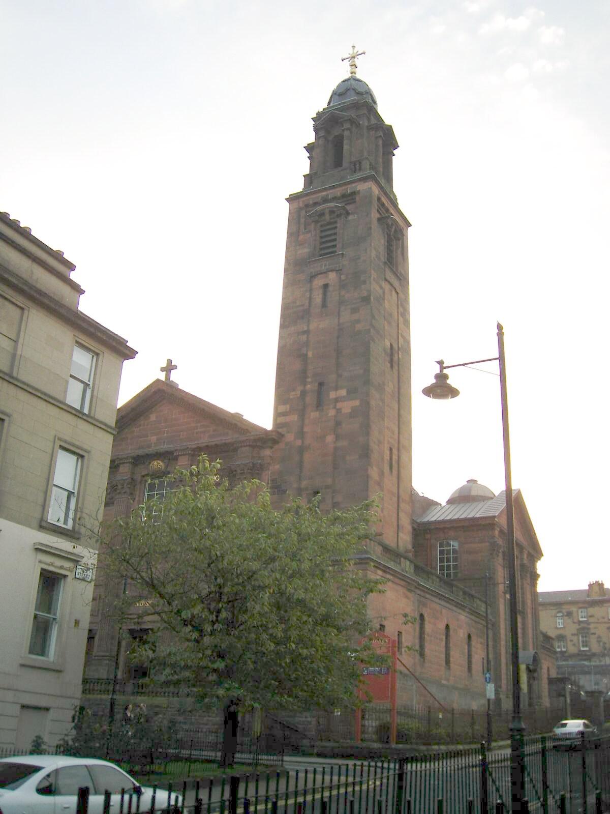 Historic church with a tall clock tower, surrounded by trees and neighboring buildings, under a soft, cloudy sky.