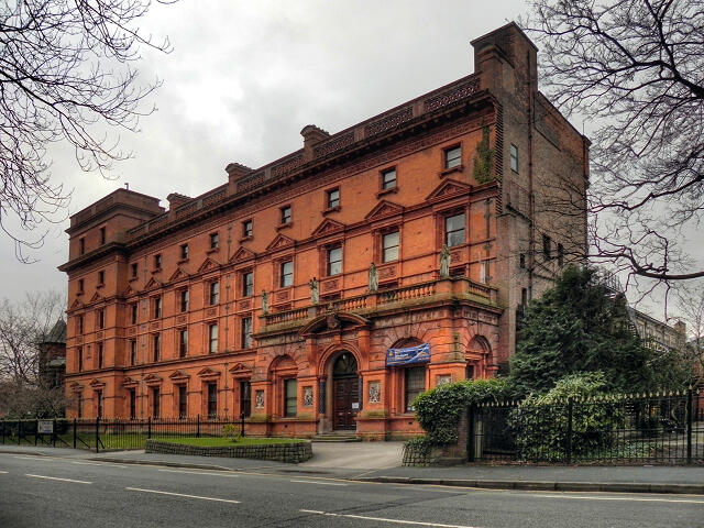 Victorian-style red brick building with ornate architectural details, surrounded by greenery and set against a cloudy sky.