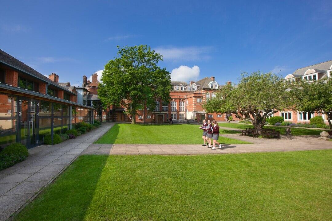 Three students walk on a sunny day across a manicured green lawn surrounded by elegant red brick buildings and trees.