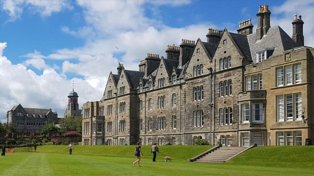 Historic stone buildings with ornate rooftops, set against a blue sky, with people walking on a lush green lawn.