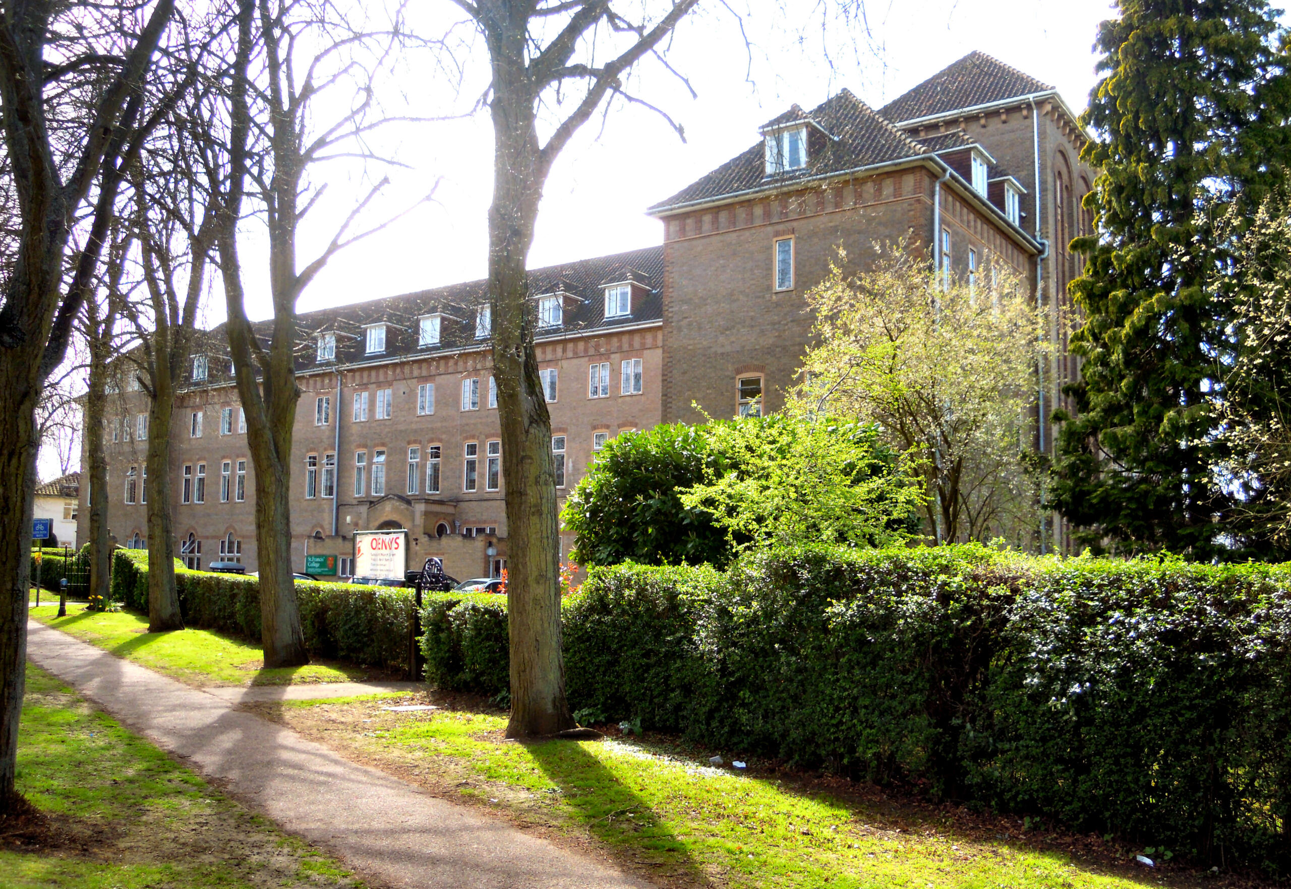 A large brick building surrounded by trees and hedges, with a pathway leading to the entrance. Sunlight brightens the scene.
