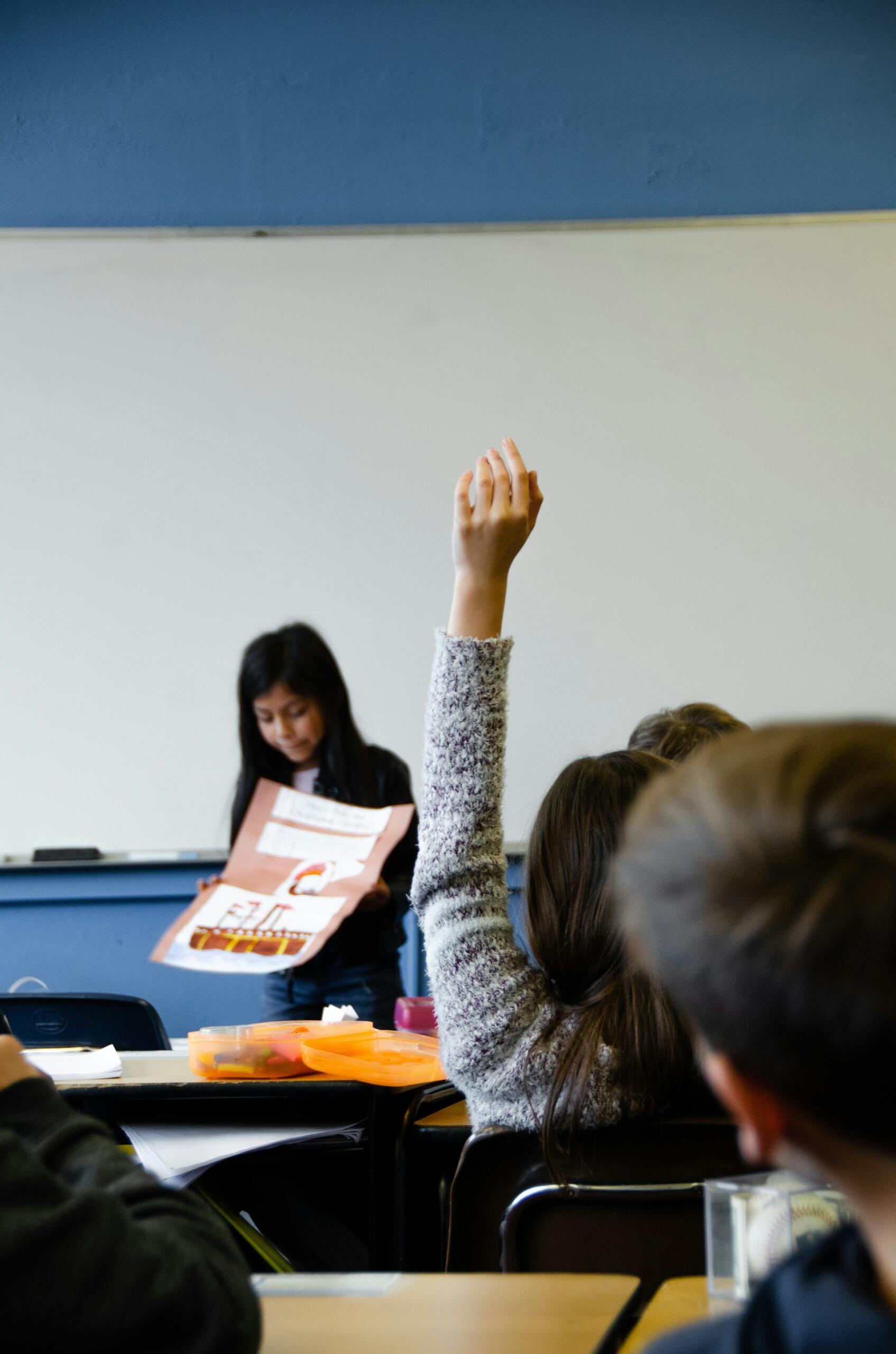 A student raises a hand in class while another presents a poster in front of a whiteboard. School supplies are on the desks.