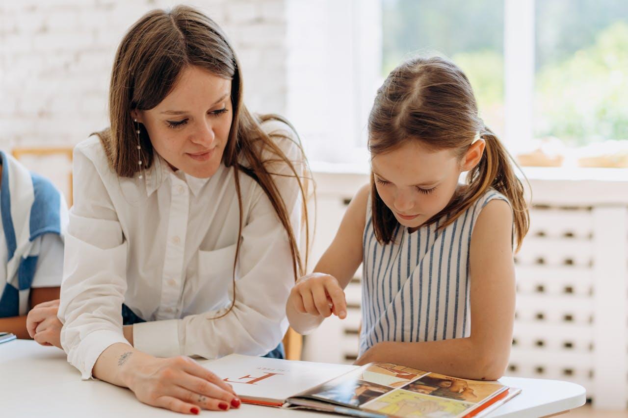 A woman and a girl are engaged in reading together, examining a colorful book at a table in a bright, sunlit room.