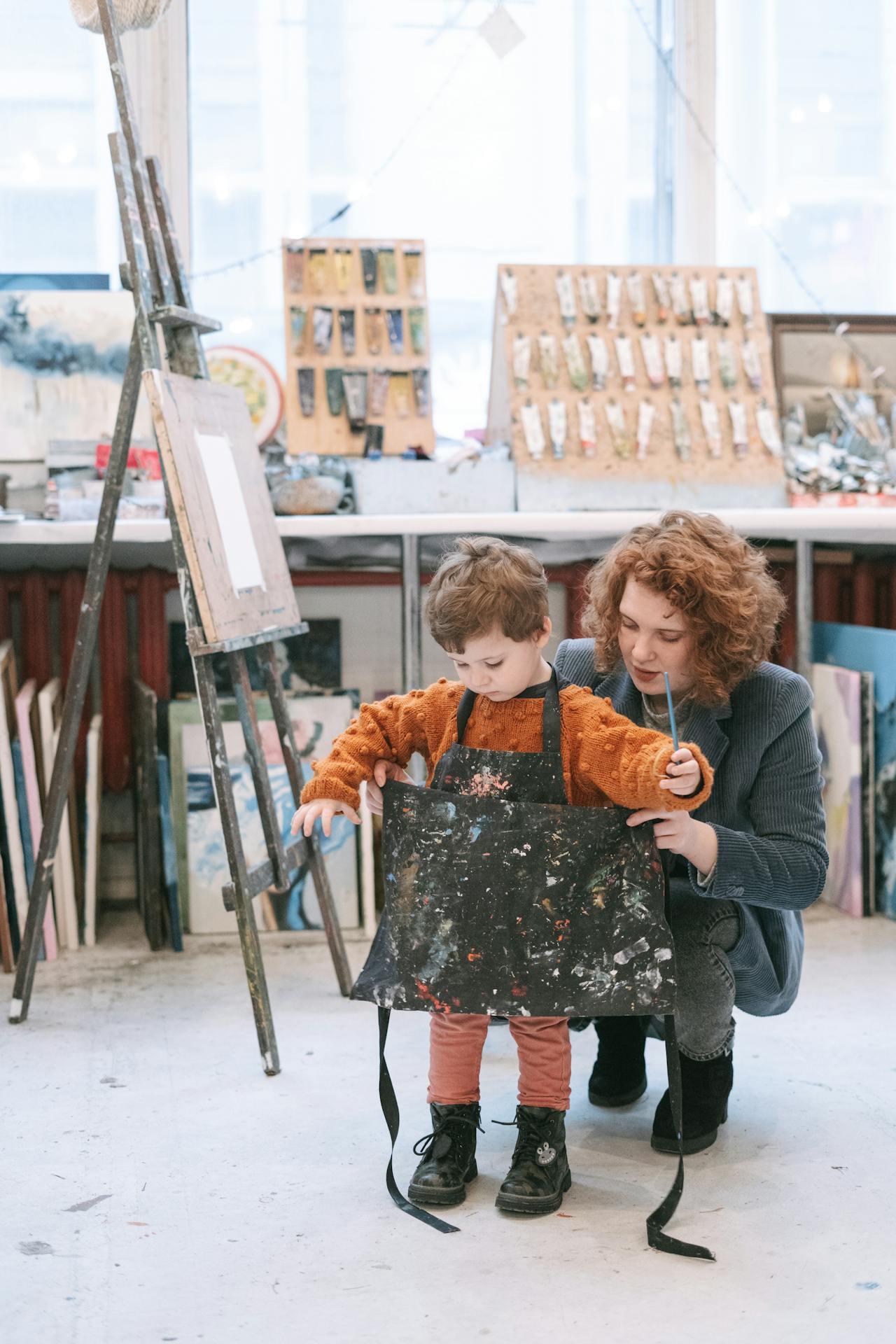 A child in a paint-splattered apron stands in an art studio with their caregiver, surrounded by easels and art supplies.