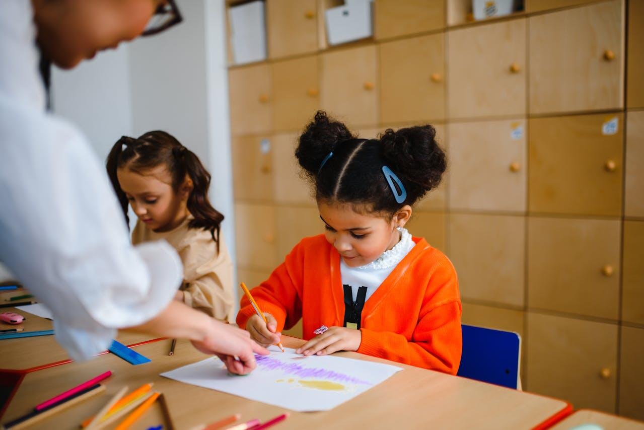 A young girl in an orange sweater colors on paper with crayons at a table, while another child watches and a hand points to the drawing.