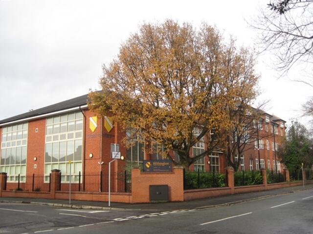 View of a brick school building with large windows, complemented by a tall tree with autumn leaves and a street sign in front.