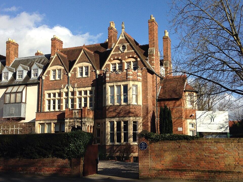 Historic brick building of Wychwood School with multiple chimneys, surrounded by trees and a sign at the entrance.