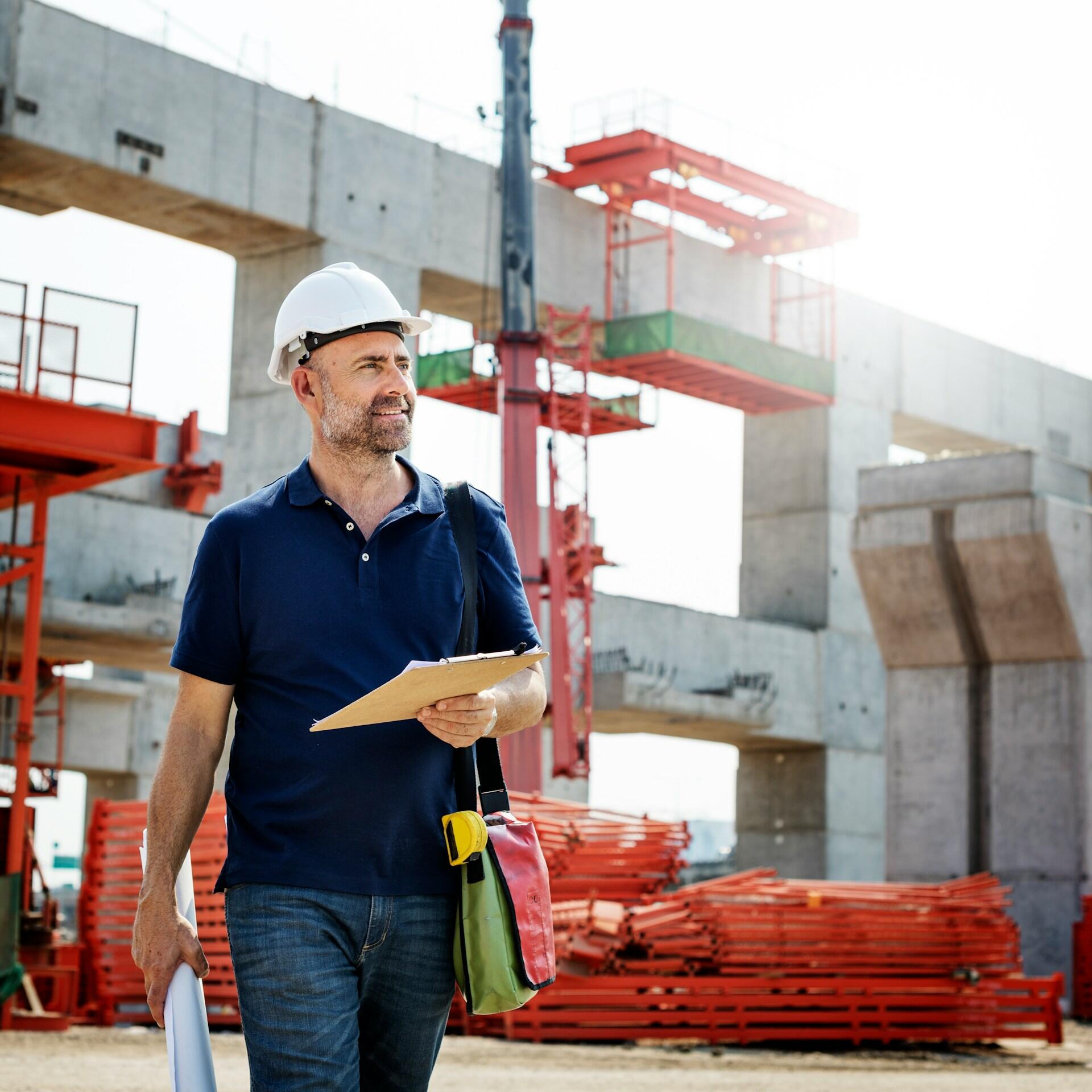 A man wearing a hardhat carrying a clipboard on a construction site. 