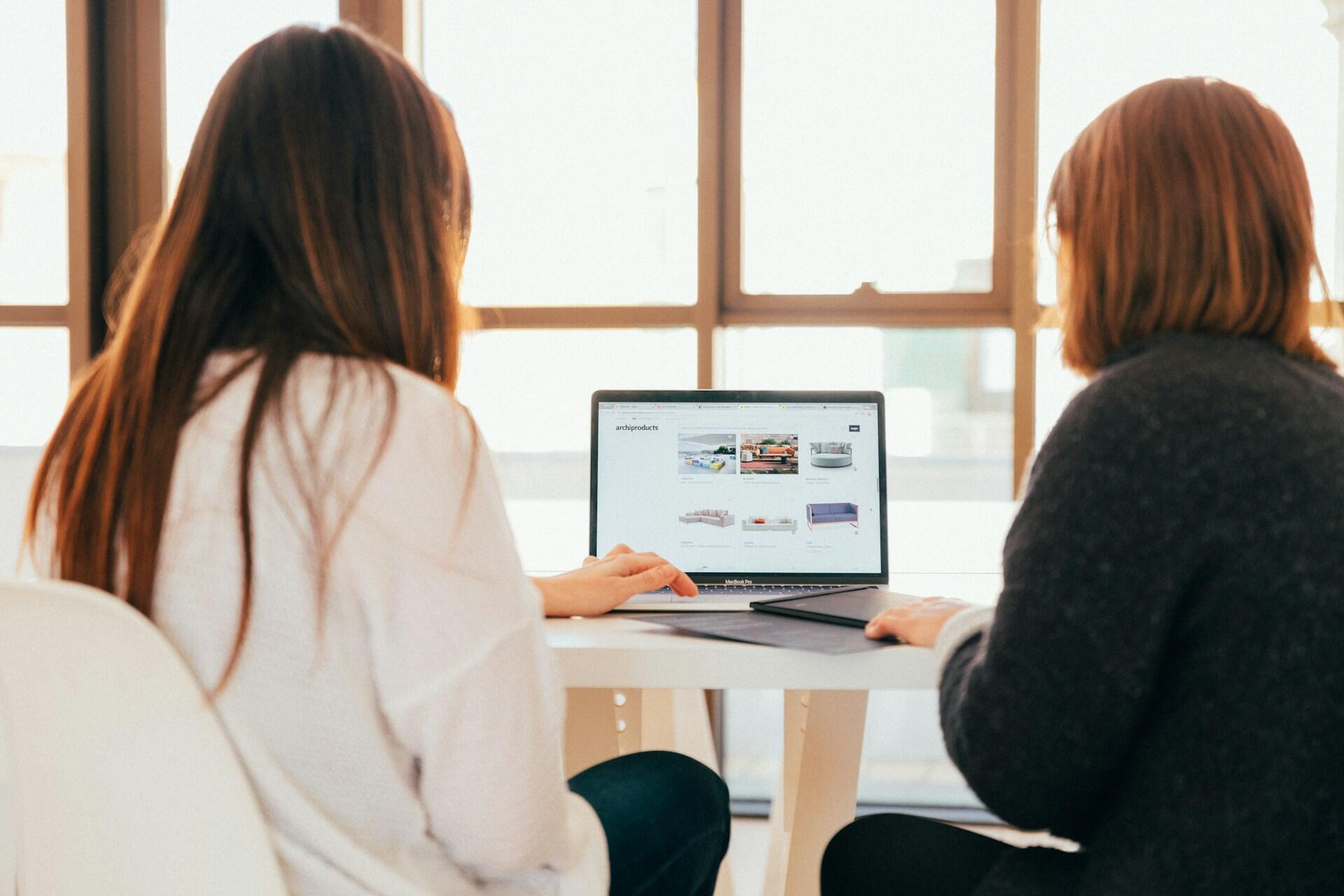 Two women sit at a computer in front of a large window. 