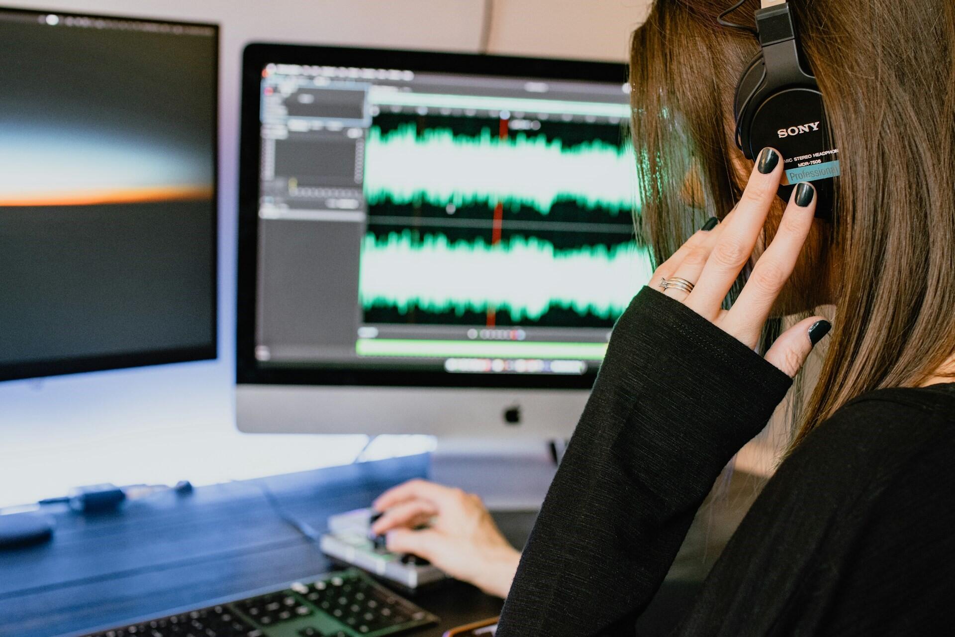 Person wearing headphones editing audio on a computer with waveform displayed on screen.