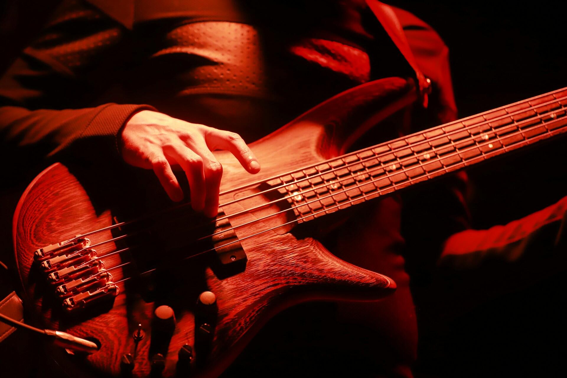 Close-up of a musician’s hand playing a bass guitar under red stage lighting.