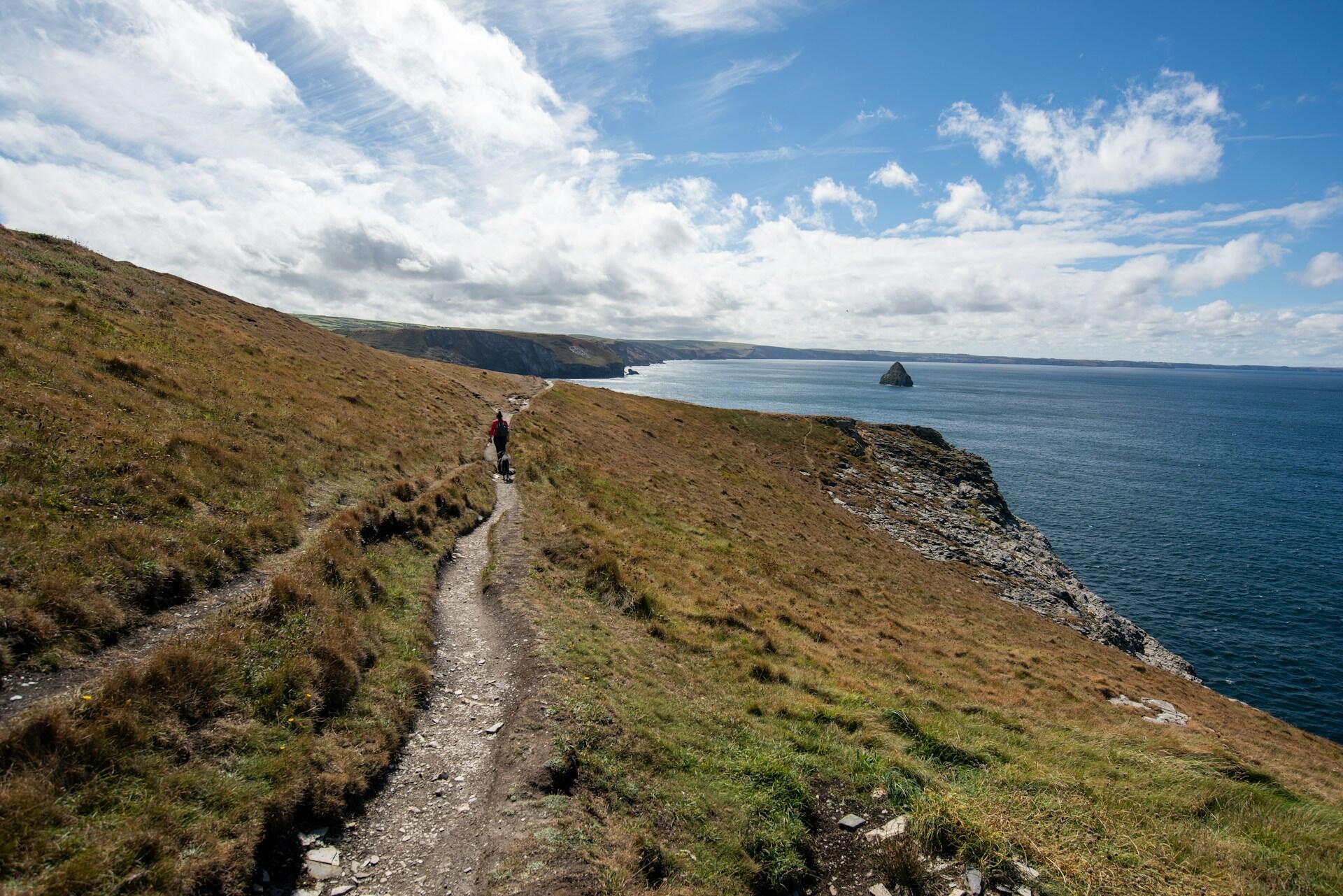 A hiker walks a dusty trail along a grassy cliff, overlooking a tranquil sea and rocky outcrop under a blue sky with scattered clouds.
