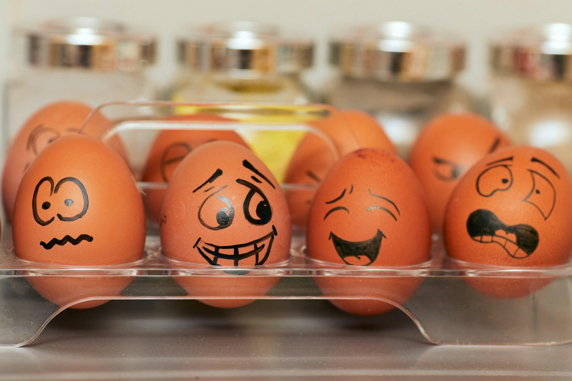A tray of brown eggs with various drawn facial expressions, including happy, confused, and surprised, against a neutral background.