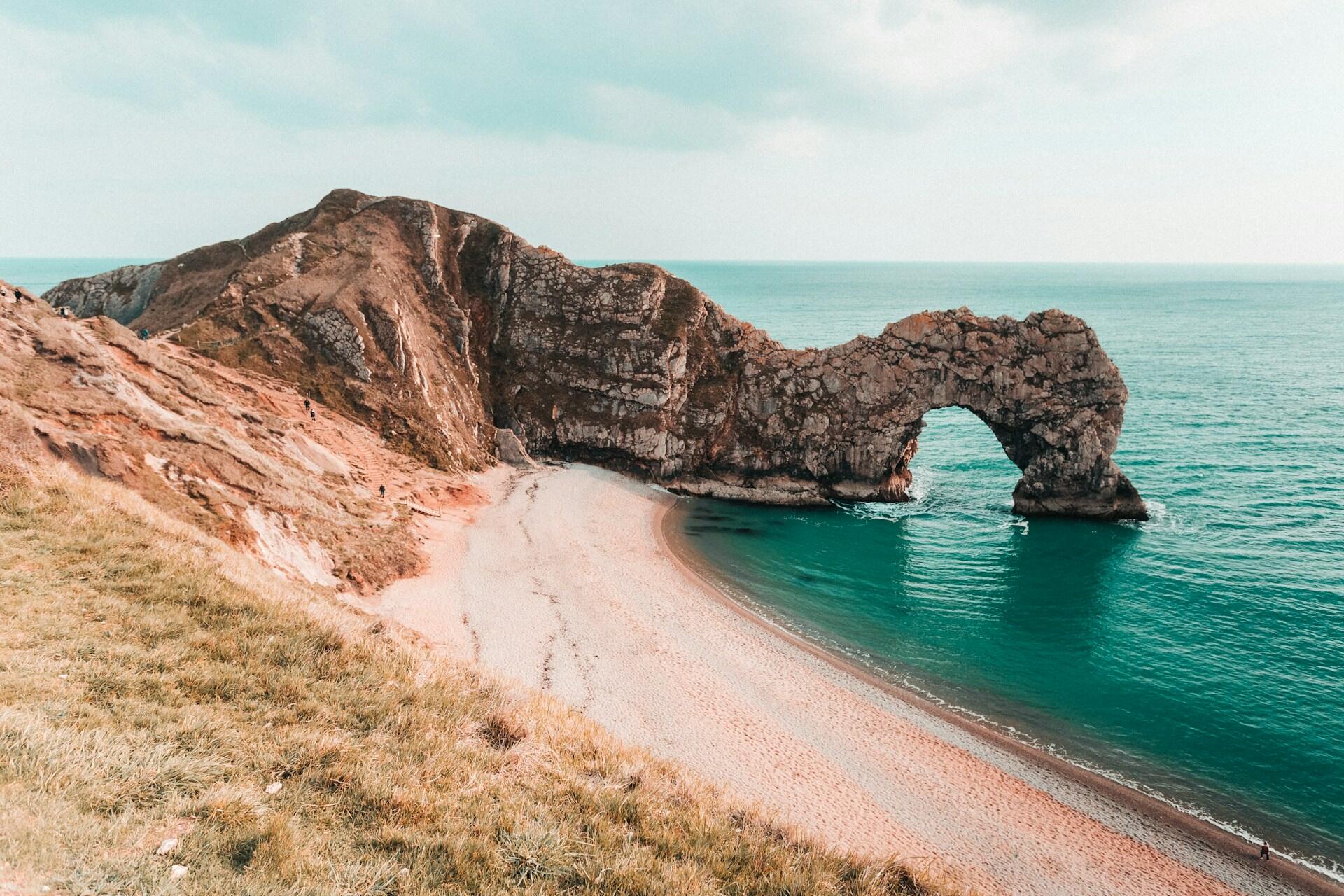 A panoramic view of Durdle Door, a natural limestone arch by the beach, with turquoise waters and grassy cliffs in the background.