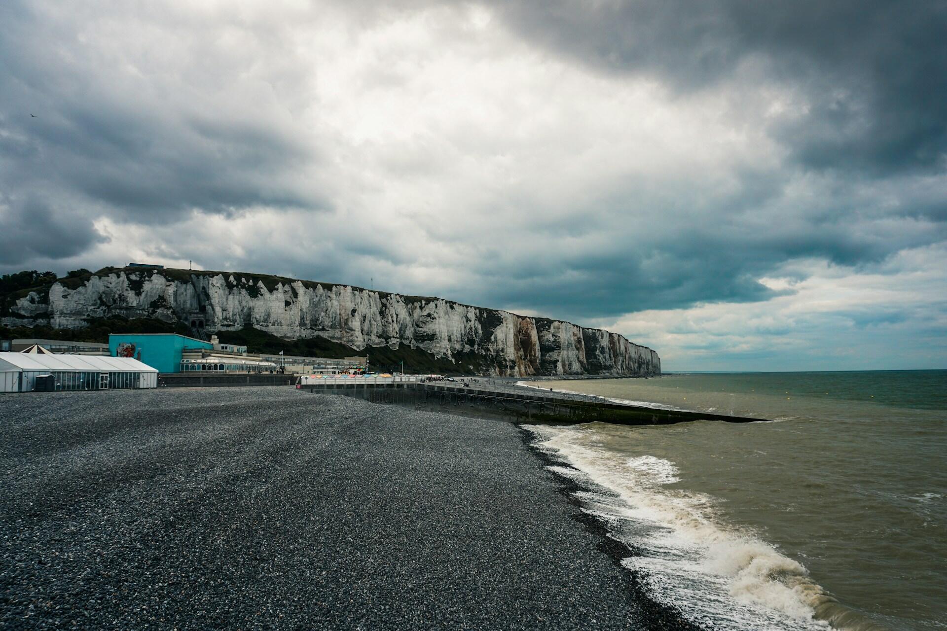 A rocky beach curves along the shore beneath dramatic gray cliffs, with stormy skies overhead and a hint of a coastal promenade in the distance.