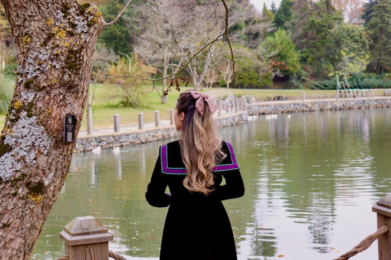 A woman with long, wavy hair and a pink bow stands by a tranquil pond, surrounded by greenery and trees.