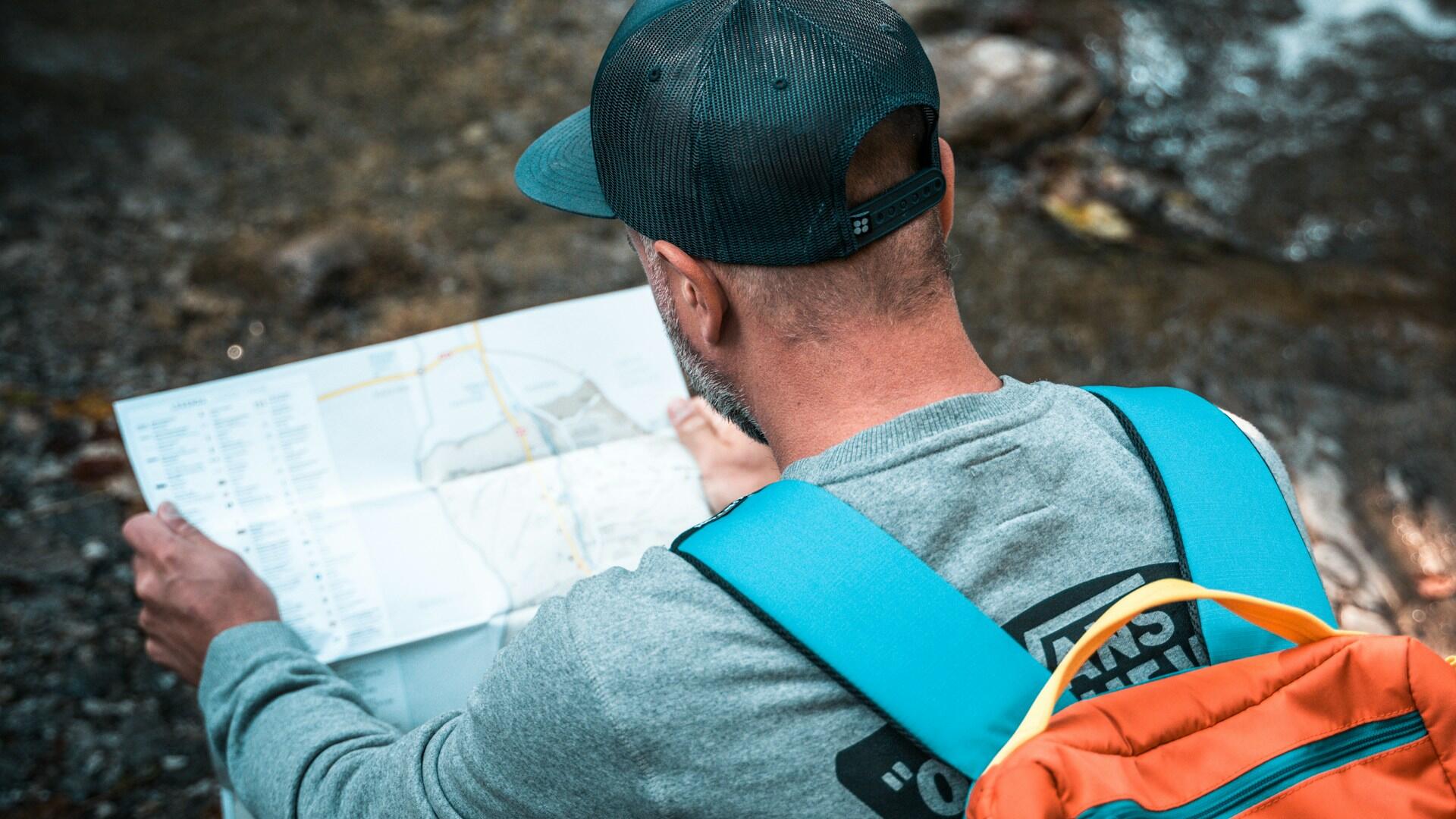 A person with a cap and gray sweatshirt studies a map by a rocky stream, carrying a colorful backpack.