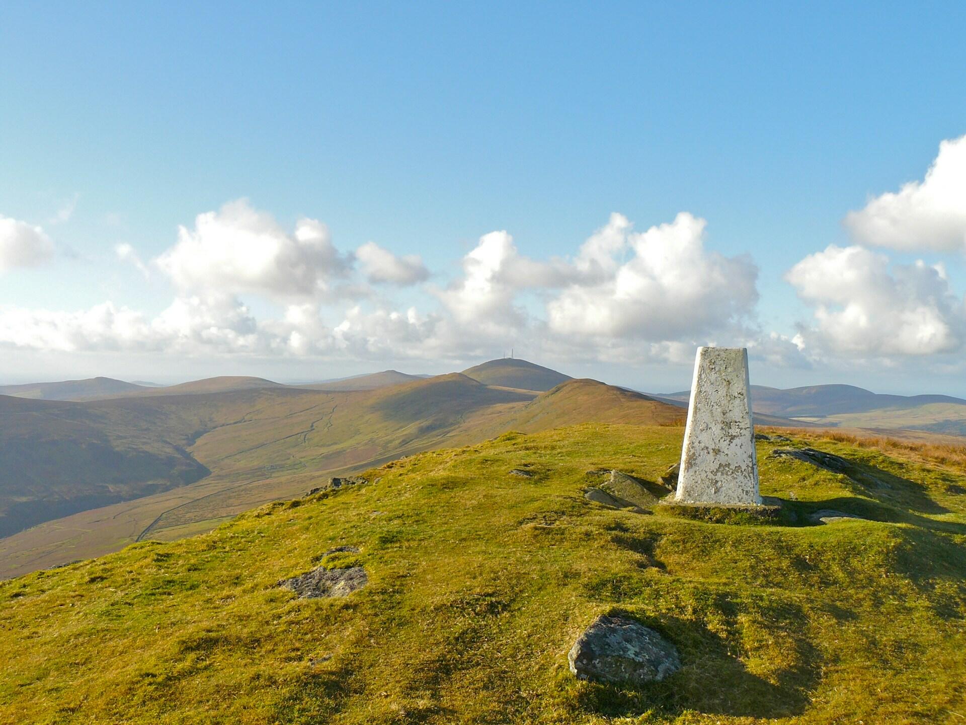 A white stone trig point stands on a green hill, overlooking rolling mountains under a blue sky with scattered clouds.