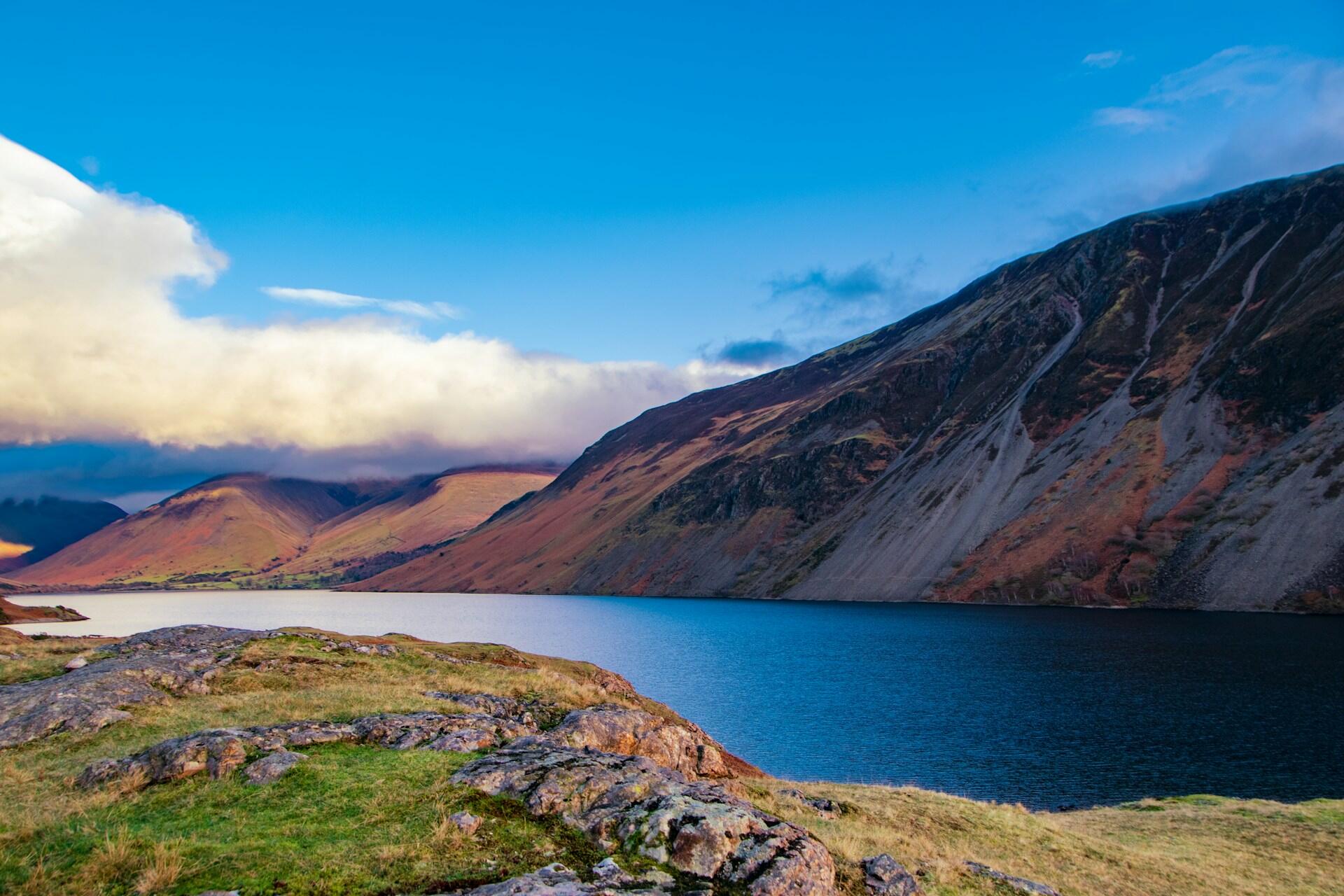 Scenic view of a serene lake surrounded by rugged hills under a clear blue sky with scattered clouds.