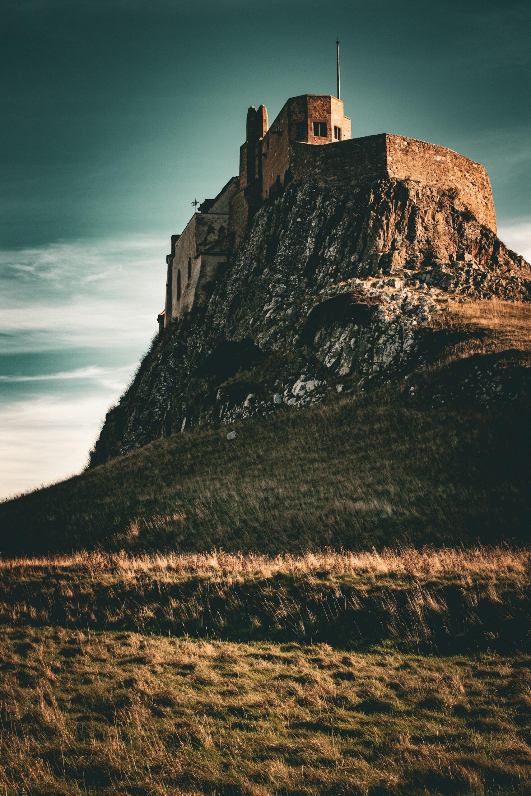 A rugged castle atop a rocky hill, surrounded by grasslands under a moody sky, with a flagpole visible at the summit.