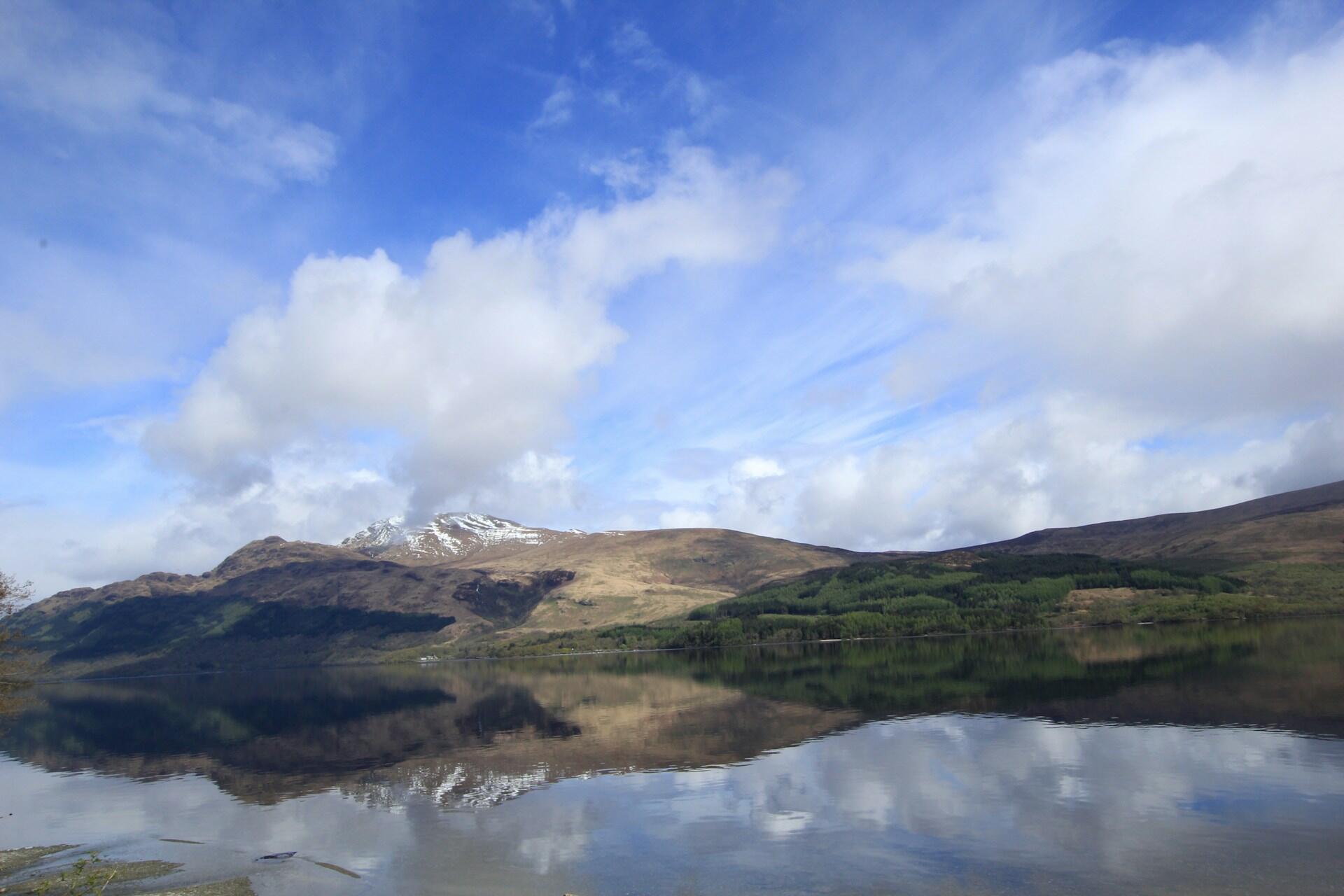 A serene landscape of a calm lake reflecting nearby mountains and a partly cloudy blue sky. 