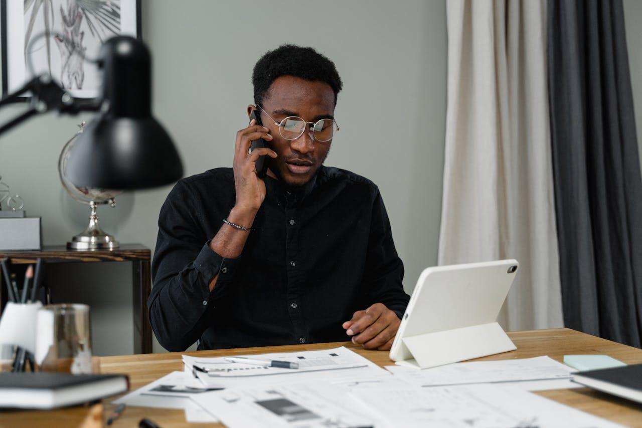 A person in a black shirt talks on the phone, surrounded by documents and a tablet on a wooden desk.