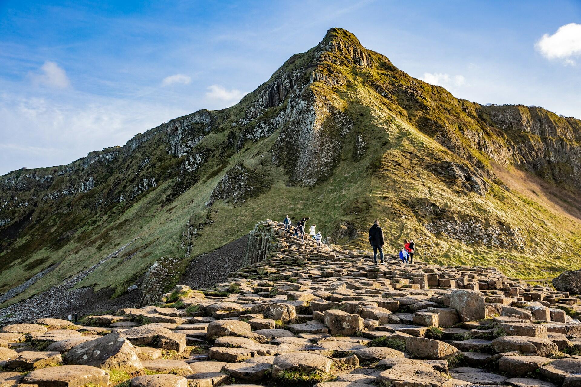People walking along a stone pathway leading up a green hillside, with rocky cliffs under a blue sky.