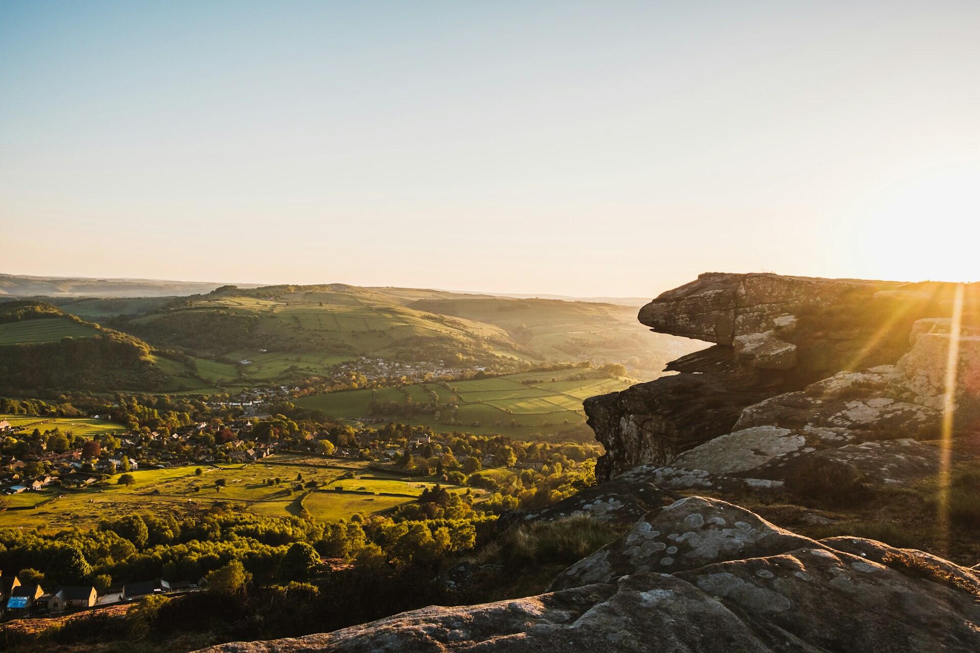 Sunrise over rolling hills, with a rocky outcrop in the foreground and a peaceful valley dotted with homes in the distance.