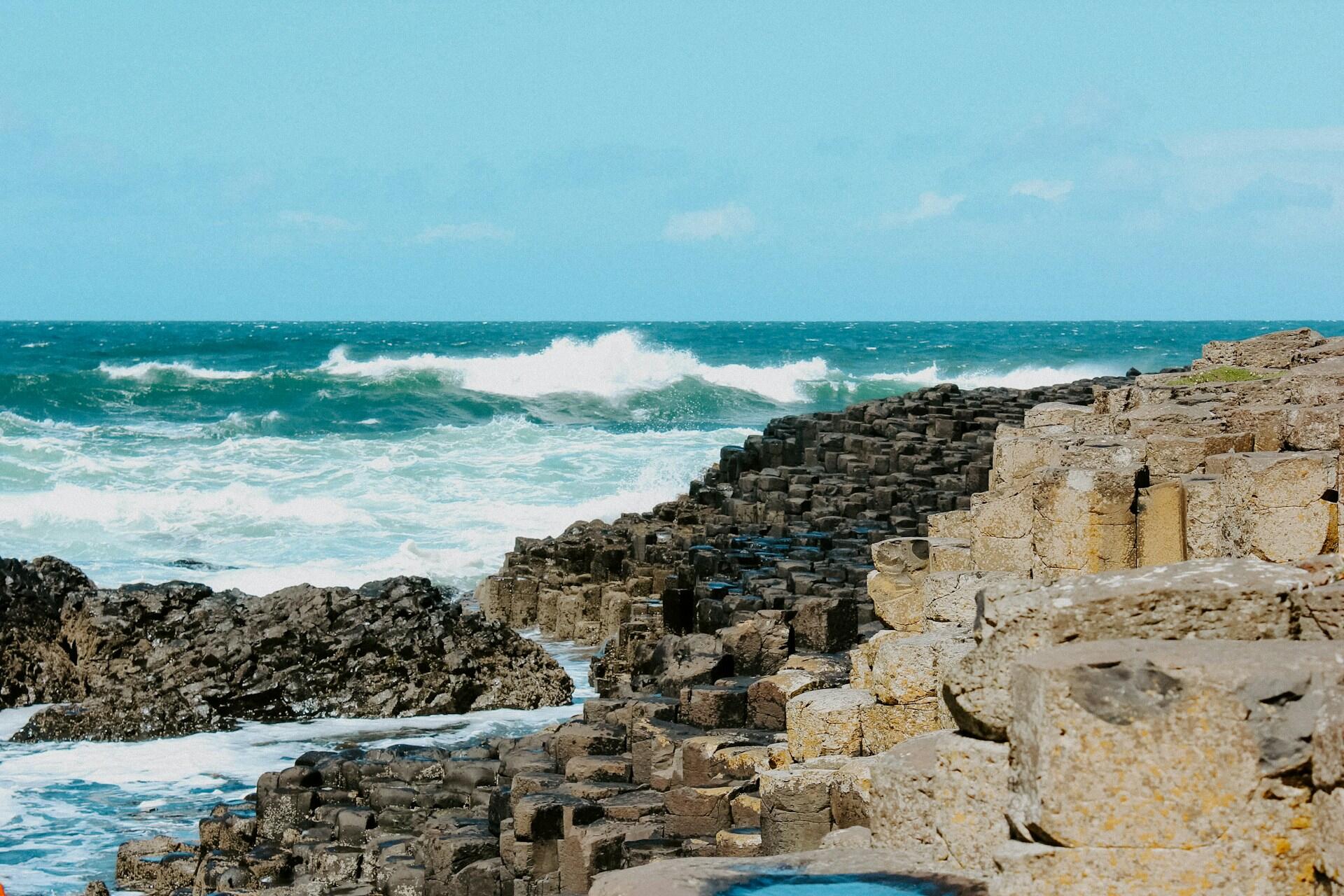 Rocky coastline with hexagonal basalt columns and crashing waves under a clear blue sky.