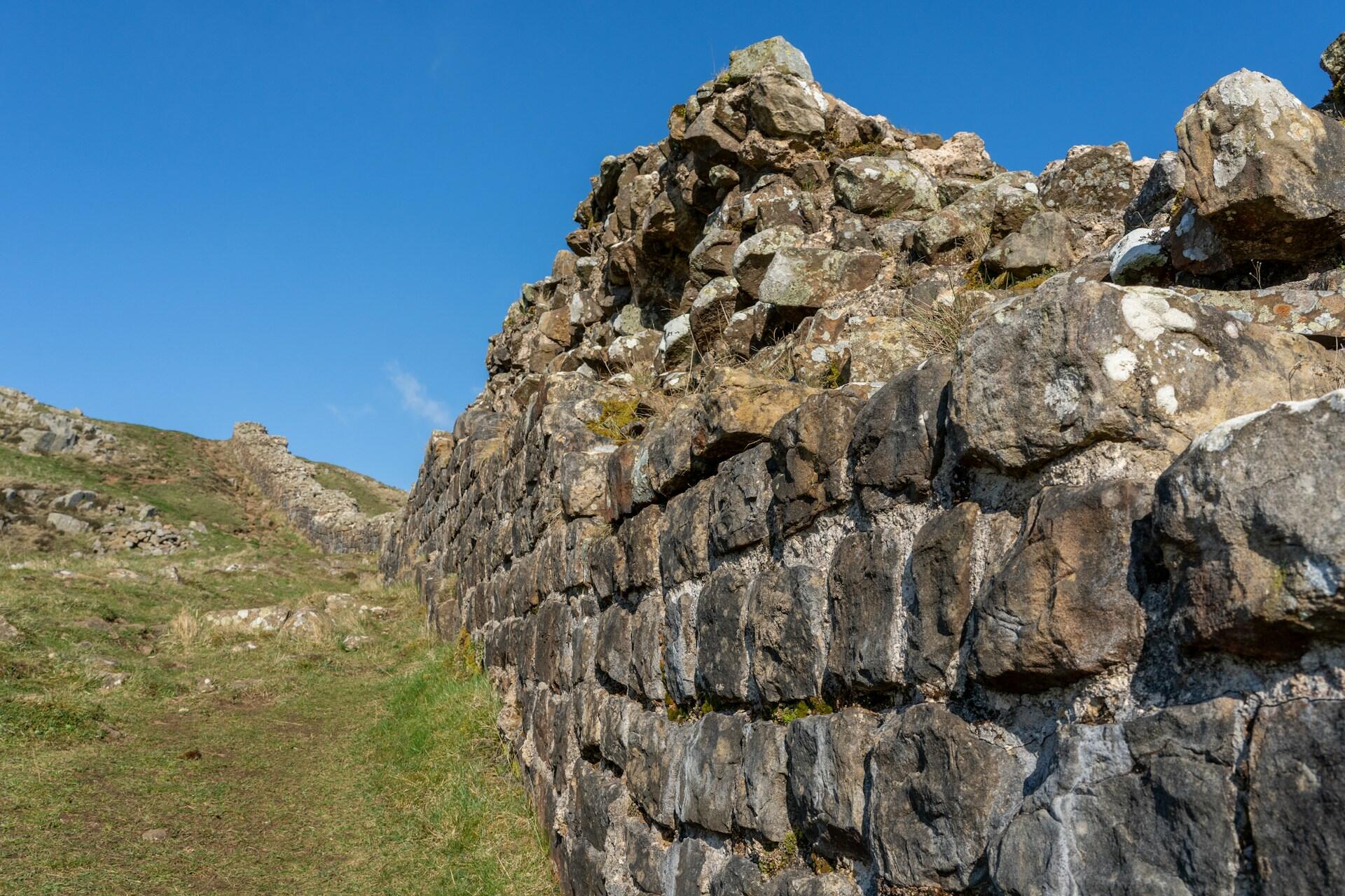 Stone wall extending into a grassy landscape with clear blue sky above, evoking an ancient, rugged feel. The scene suggests historical significance.
