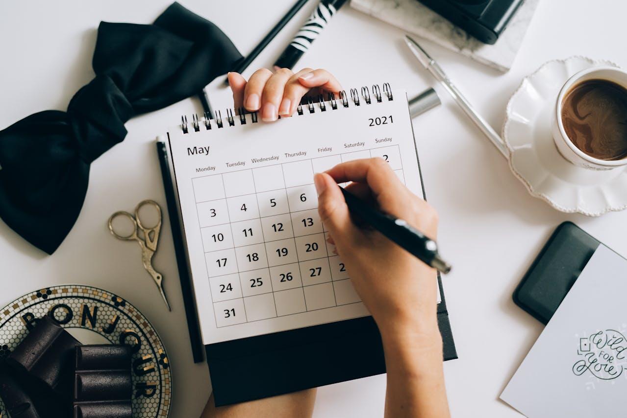 A person writes on a May 2021 calendar surrounded by scissors, chocolate, a coffee cup, and various stationery items.