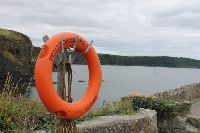 Orange lifebuoy on a stone cliffside, overlooking a calm sea under cloudy skies. Grass grows nearby, creating a serene, coastal scene.