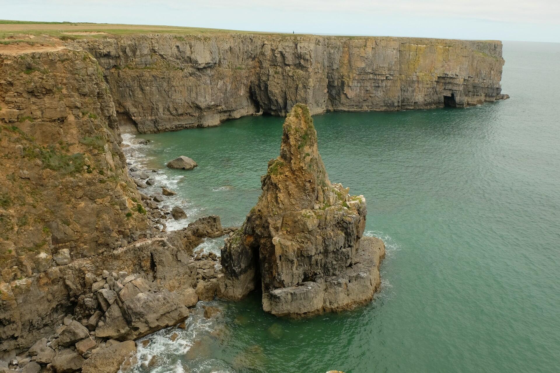 A rocky cliff juts into the turquoise sea, with a tall stone spire and gentle waves lapping at the shore under an overcast sky.