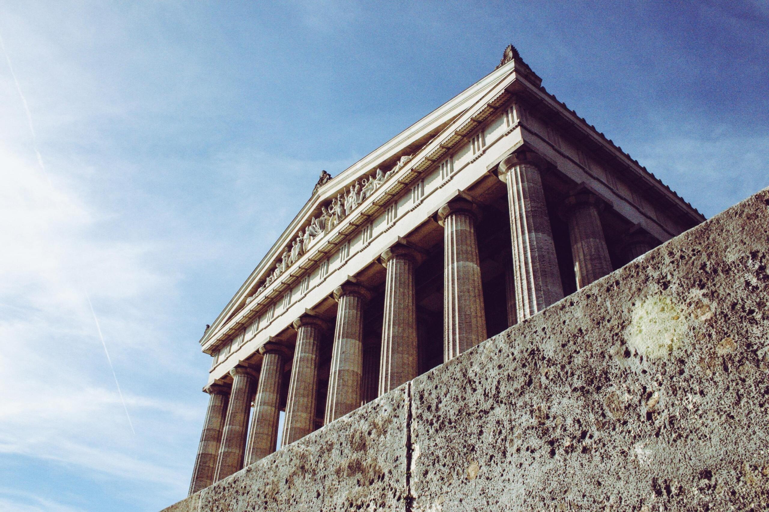 An ancient Greek building with tall stone pillars stands under bright sunlight, showcasing its classical architectural design and historic craftsmanship.