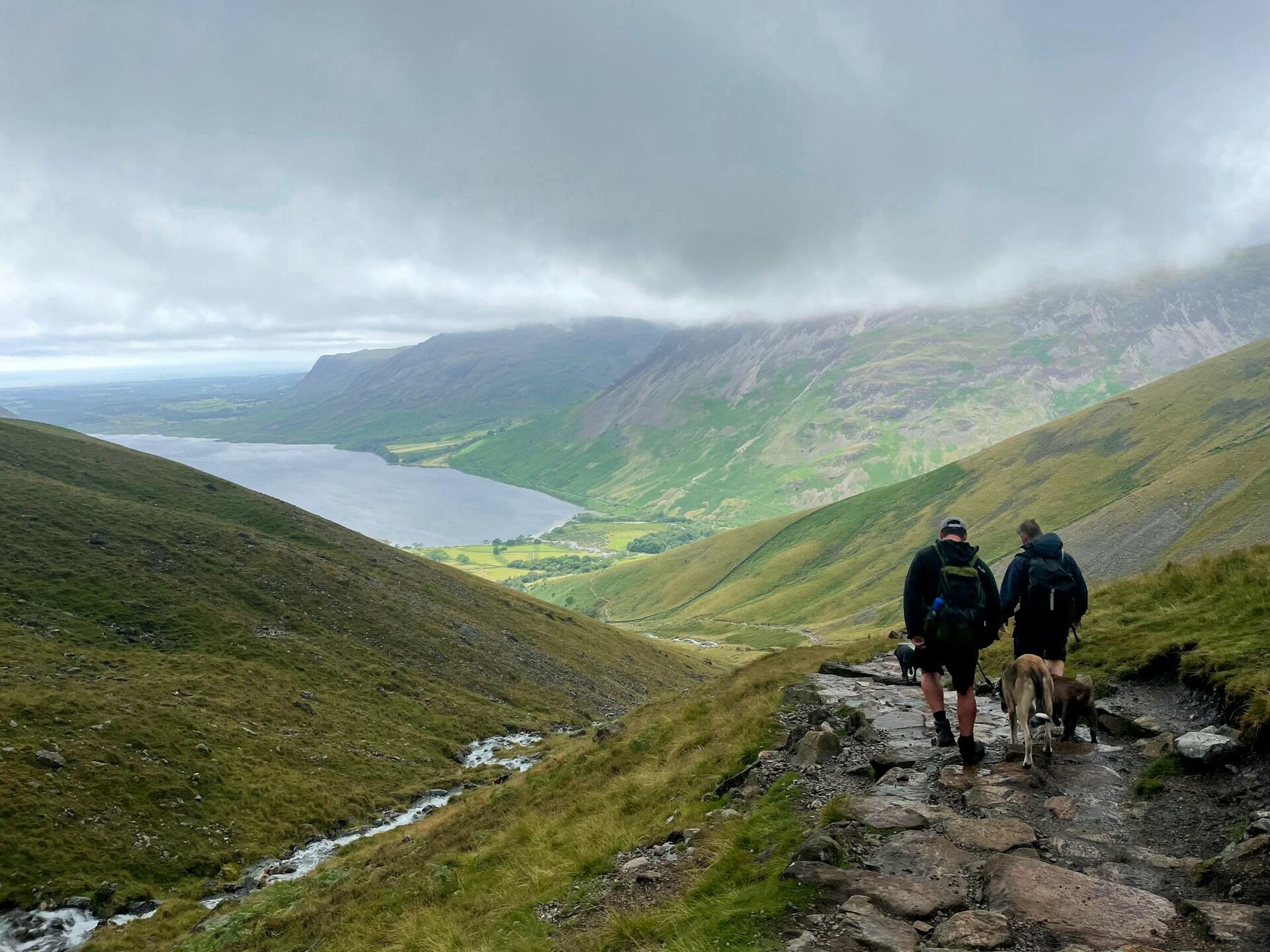 Two hikers and a dog trek along a rocky trail, overlooking a tranquil lake and lush green valleys under a cloudy sky.