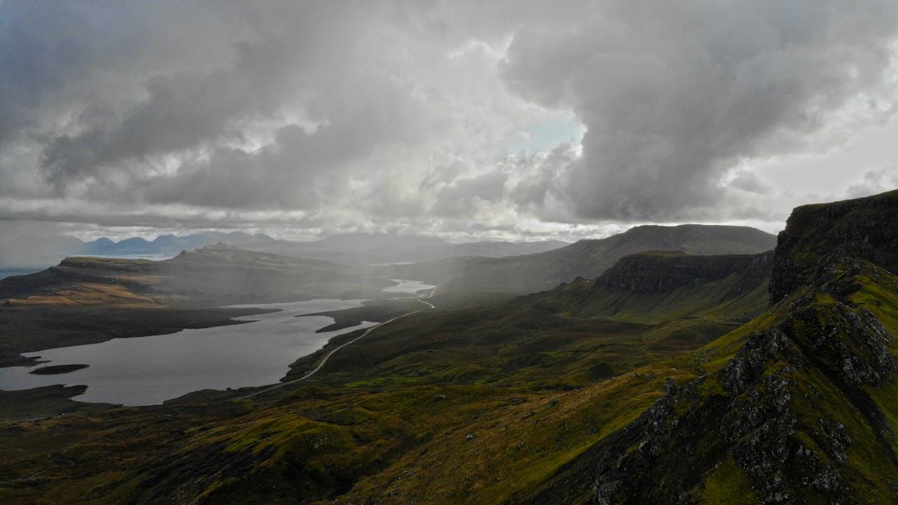 A dramatic landscape featuring a winding river, lush green hills, and dark, cloud-covered mountains under a moody sky.