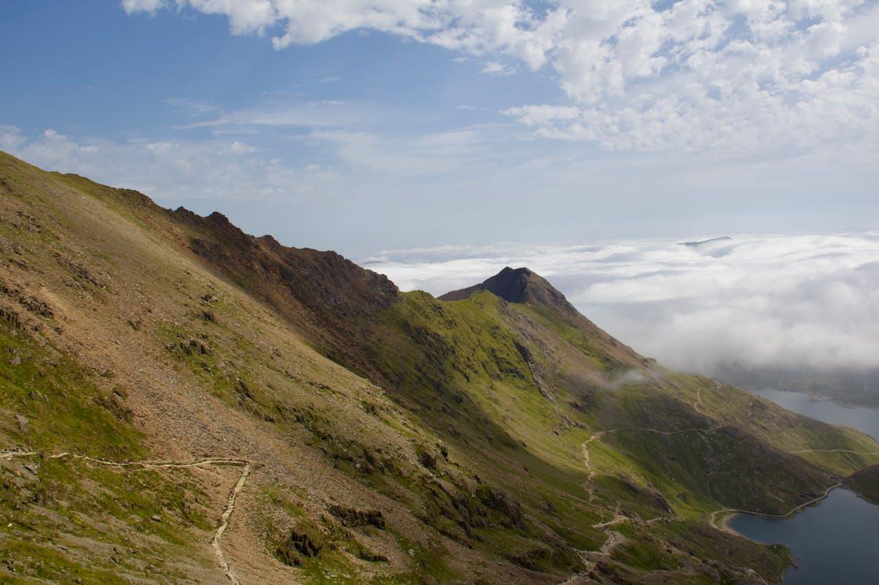 Vast mountain scenery with rocky slopes, green hills, and a lake below, shrouded in mist under a bright, cloudy sky.