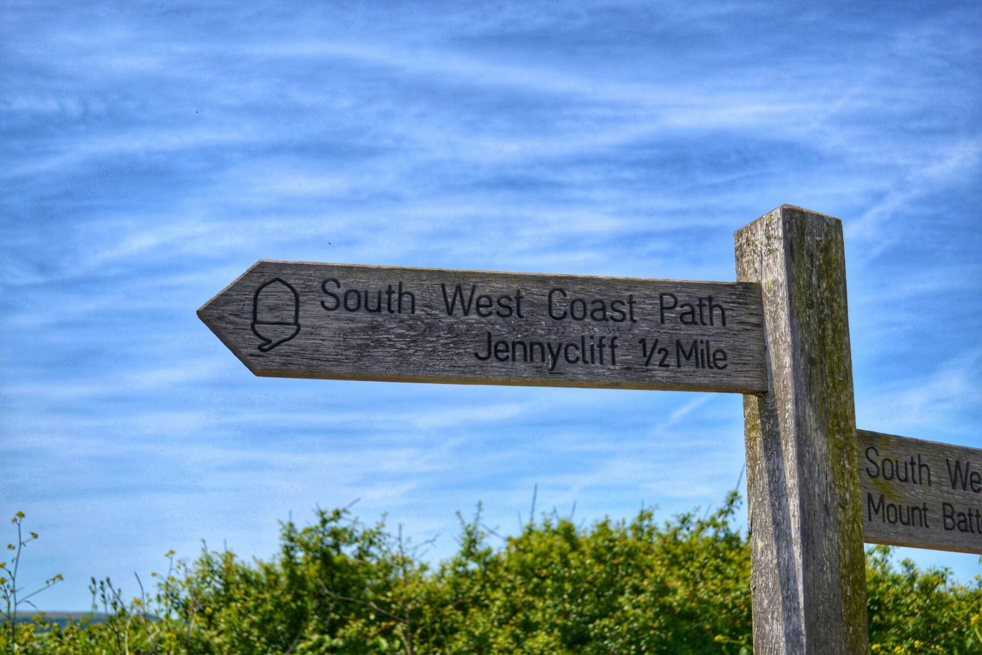 Wooden signpost pointing towards "South West Coast Path - Jennycliff ½ Mile" under a blue sky, surrounded by greenery.