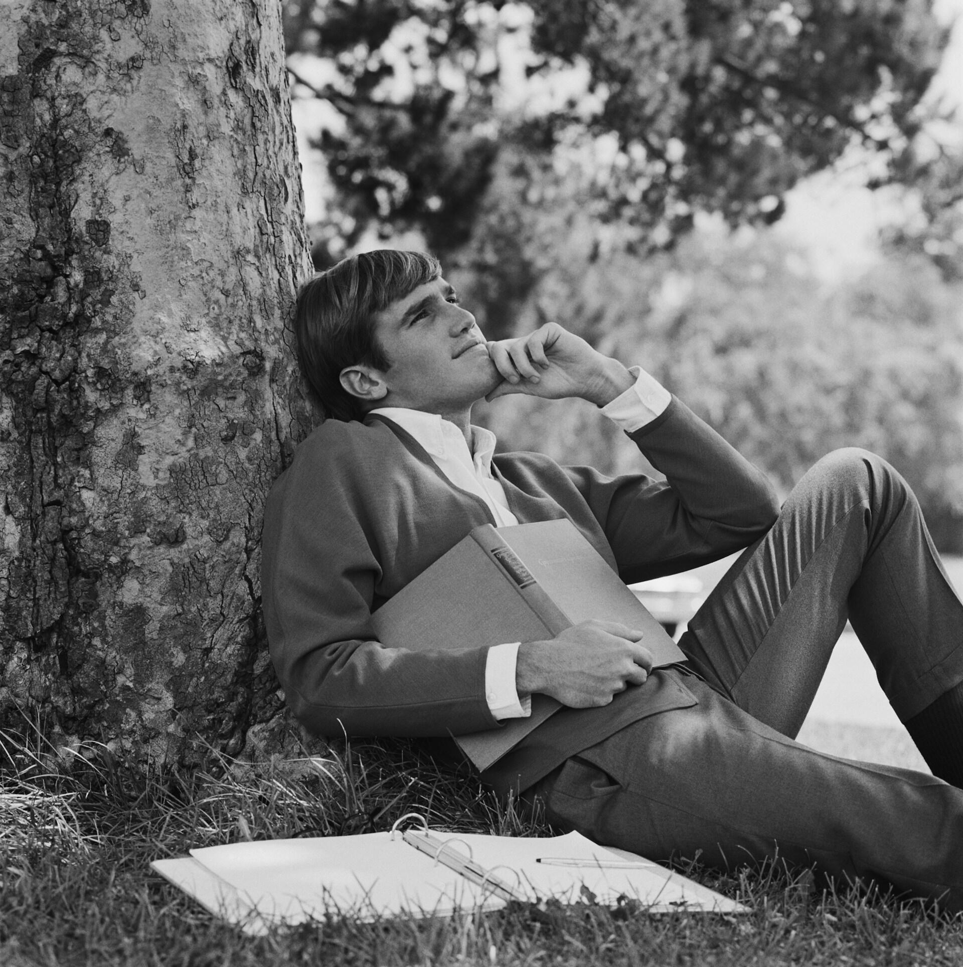 A man in a suit holding a book reclines against a tree. 