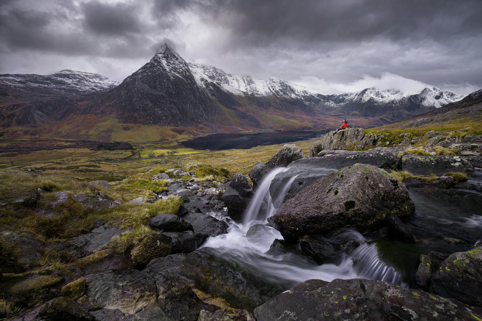 A lone figure in red sits on rocks by a waterfall, overlooking a dramatic landscape of mountains and a serene lake under cloudy skies.