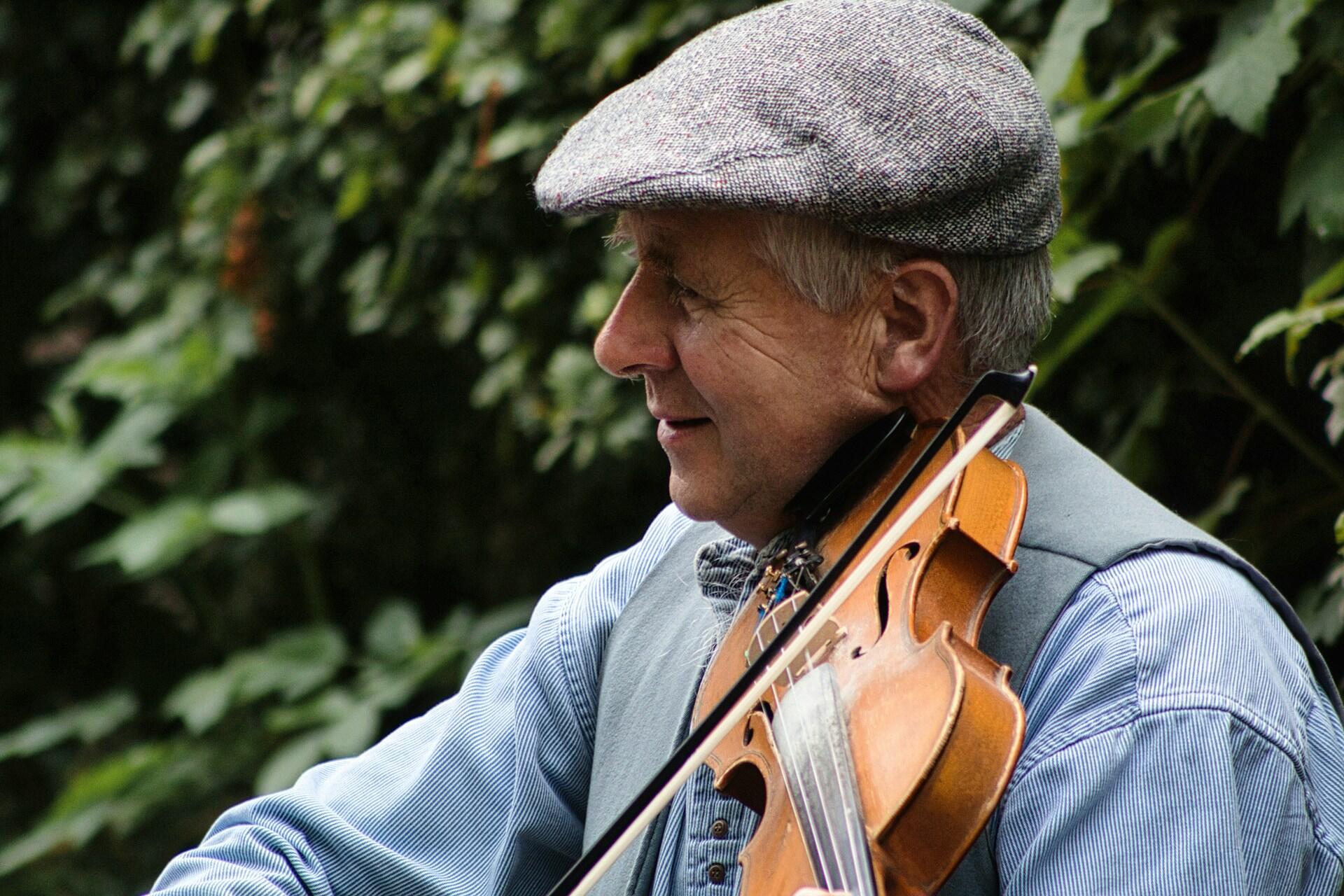 Older man wearing a flat cap playing a violin outdoors among trees.