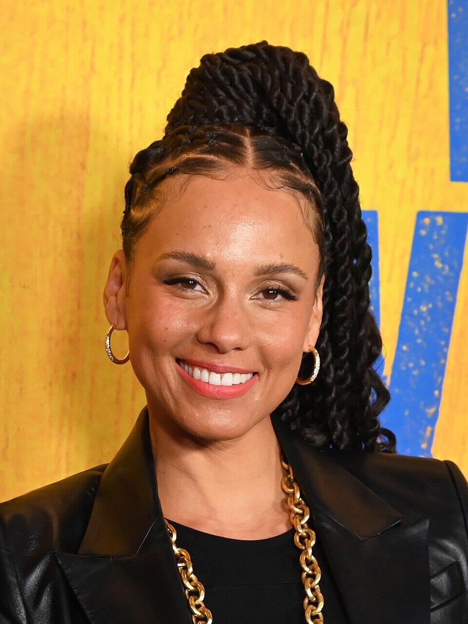 A person with stylish braided hair, wearing a black blazer and a chunky gold chain, stands against a vibrant yellow background.